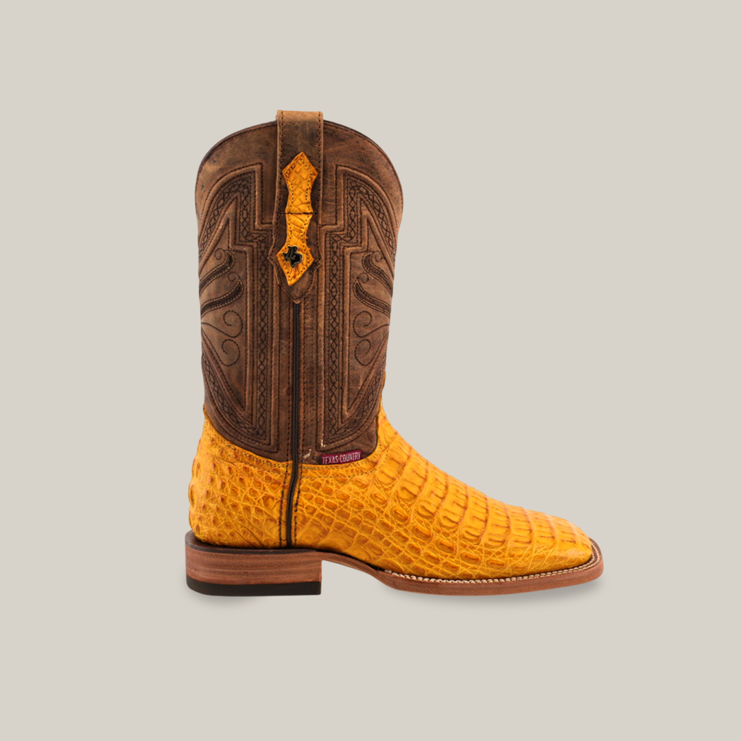 The Exotic Caiman Horn Back - Buttercup-Square Toe boot features a side view with a brown, intricately stitched upper and bright yellow caiman leather-textured foot. The luxurious design includes a wooden heel and dark sole, capturing the cowboy lifestyle against a plain backdrop.