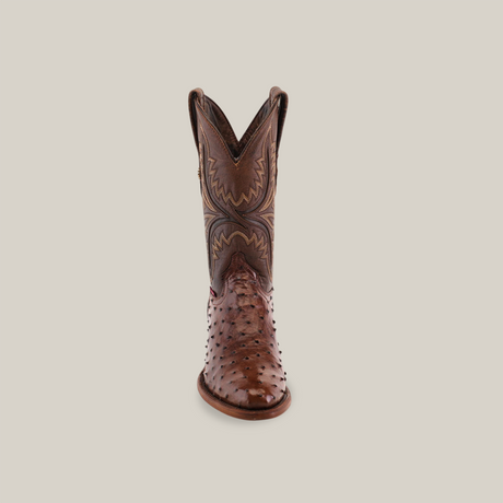 The Exotic Full Quilt Ostrich - Brown - Round Toe boot displays exquisite leather artistry with its intricate embroidery on the tall shaft. Handcrafted to perfection, its textured surface contrasts against a plain light-colored background.