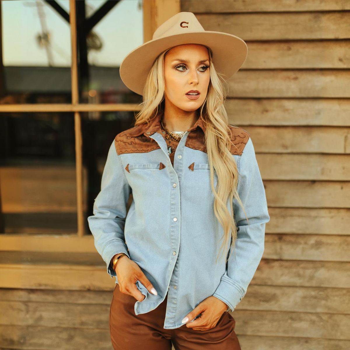 A woman with long blonde hair, wearing a beige wide-brimmed hat and an LB Denim Camel Suede Pearl Snap TO718 shirt, stands in front of a rustic wooden building. She looks to the side with one hand tucked into the pocket of her brown pants.