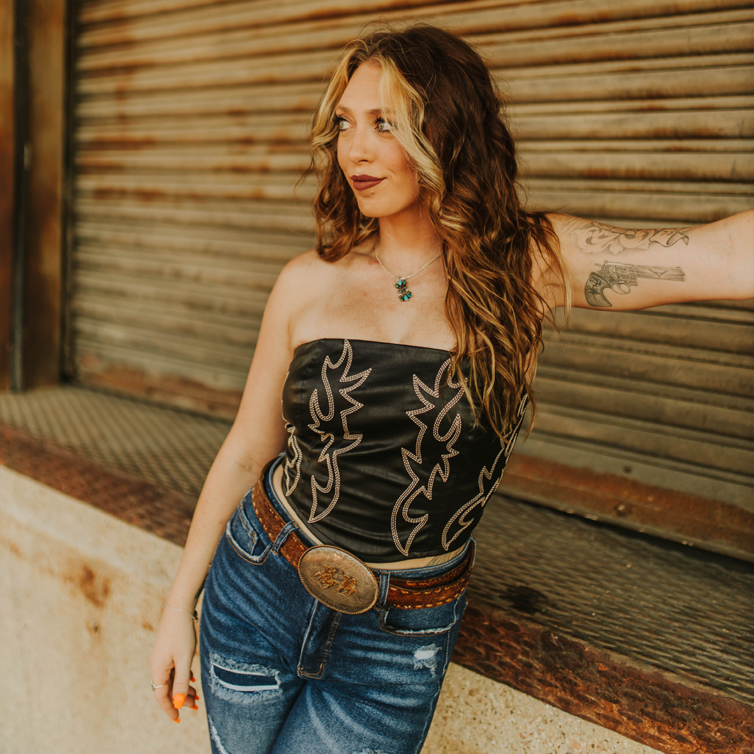 A woman with long, wavy hair and a tattoo on her arm leans against a metal shutter. She wears an LB Black Leather Corset Western Stitch (TO720-BLK) with intricate designs, paired with jeans and a western stitch belt, gazing to the side while standing on the rustic concrete.