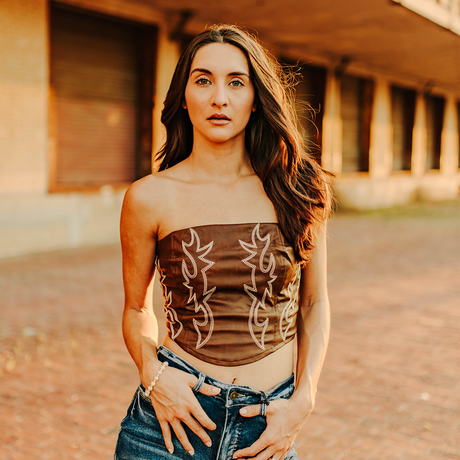 A woman with long brown hair stands outdoors on a brick path, wearing the LB Brown Leather Corset Western Stitch TO720-BRW and blue jeans. The background features a building with large windows and a rustic look.