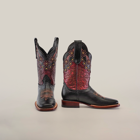 A pair of Grecas Black boots with ornate handcrafted black and burgundy leather, intricate floral embroidery, and decorative stitching. One boot stands upright to highlight the square toe design; the other is tilted to display the craftsmanship