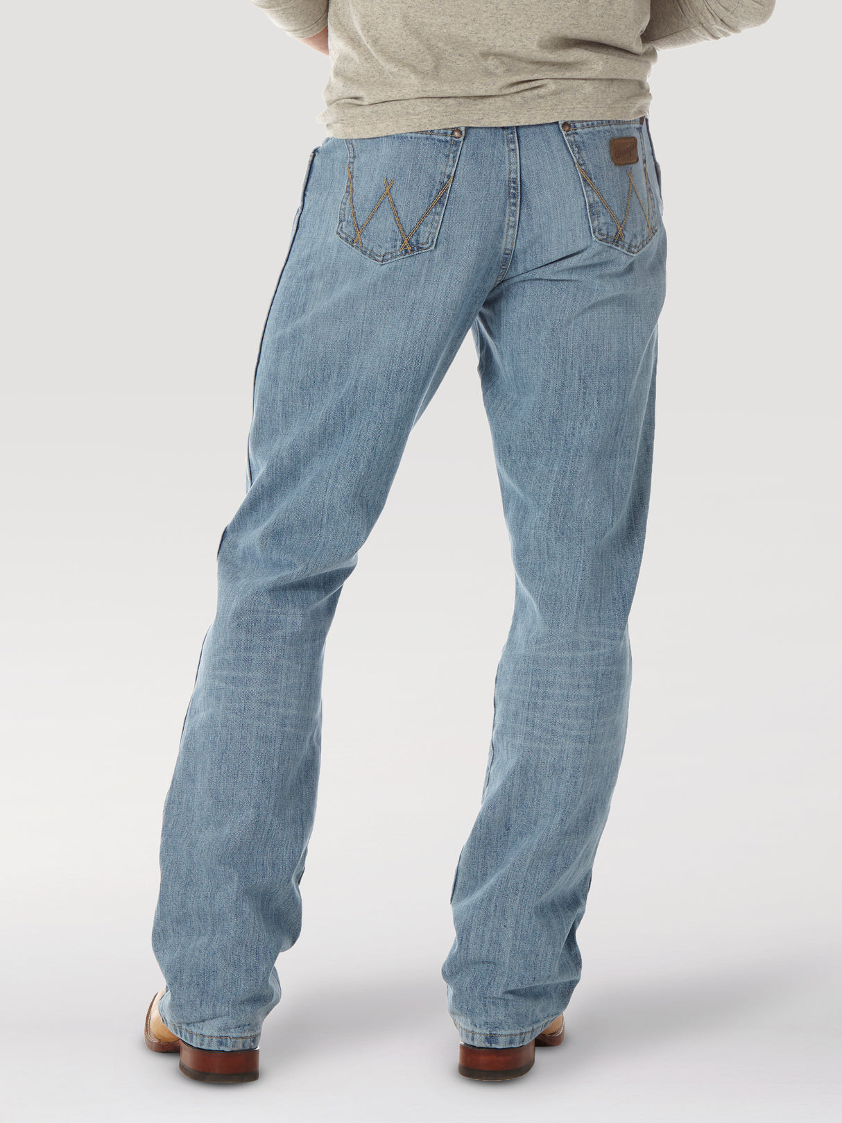 A rear view shows a person wearing Mens Wrangler Retro Relaxed Fit Bootcut Jeans - WRT20CR, in light blue, paired with a gray long-sleeve shirt. The jeans display back pockets with decorative stitching and a leather patch on the waistband, set against a light background.