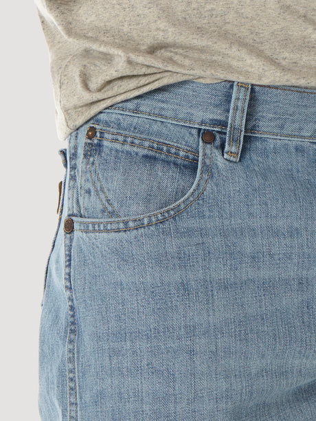 A close-up shows a person wearing the Mens Wrangler Retro Relaxed Fit Bootcut Jean - WRT20CR with a gray shirt. The light blue denim features bootcut style, front pocket with copper rivets, visible stitching, and a classic, slightly worn texture.