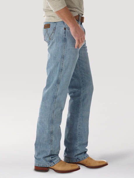 A person in light blue Mens Wrangler Retro Relaxed Fit Bootcut Jeans (WRT20CR) with a brown leather patch and subtle back pocket embroidery, paired with a brown belt, beige cowboy boots, and a light gray long-sleeve shirt, stands against a plain background.