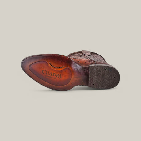 The image highlights the sole of a Moroni Chestnut Ostrich Laser & Embroidery - Round Toe boot. The textured sole displays the embossed Cuadra logo with visible wear marks. Detailed stitching and craftsmanship, with ostrich leather accents, enhance its exquisite look.