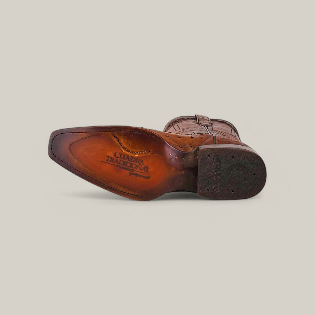 An Arauco Chocolate Ostrich CU597 cowboy boot with a narrow square toe lies showcasing the laser-engraved sole saying Cuadra Tradición and Handmade, highlighted against a light background, featuring intricate stitching.