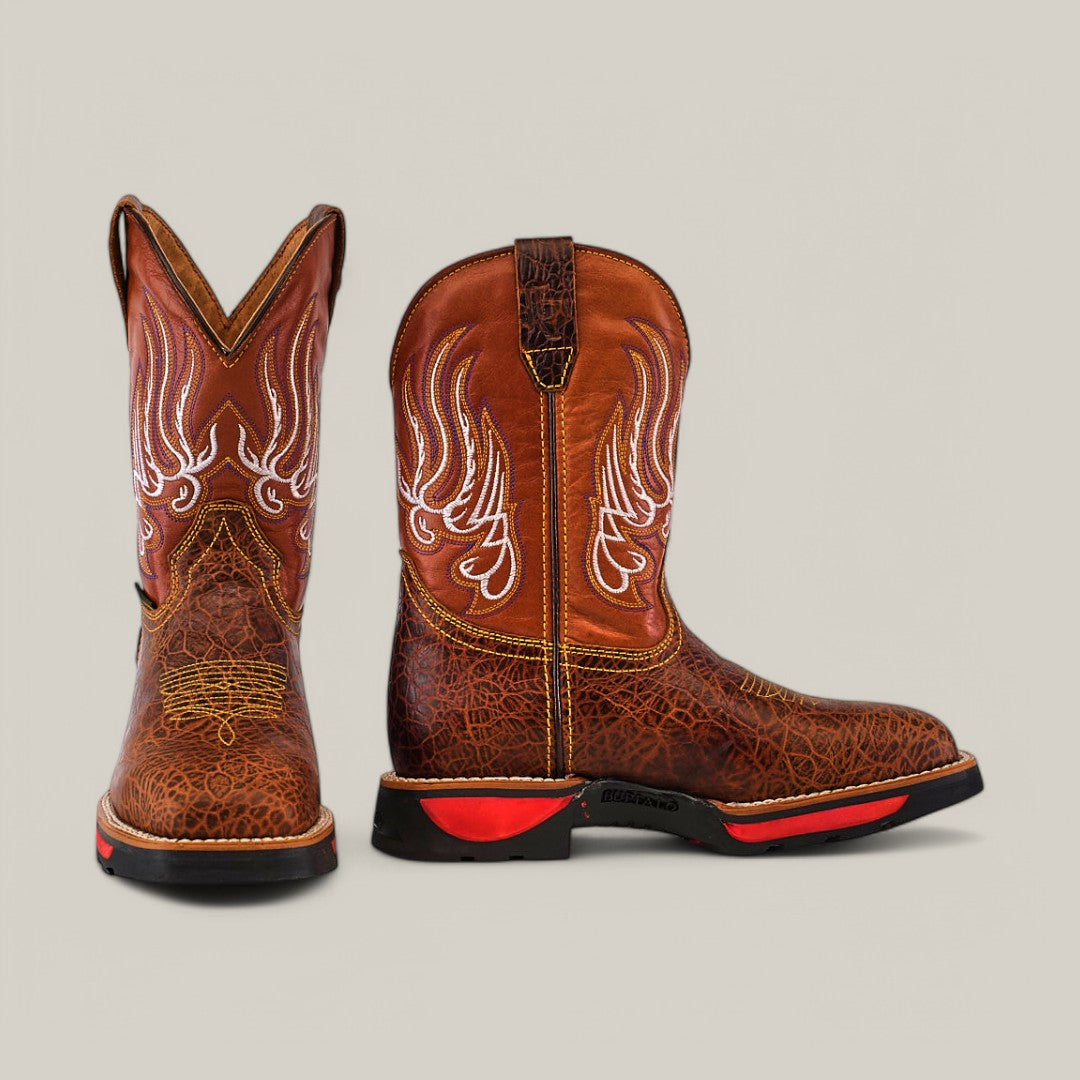 The Jurasic Honey Composite Toe - E6080 features brown cowboy boots with intricate white embroidery and striking red soles, set against a plain light backdrop. One boot faces forward while the other is in profile, highlighting the durability typical of Texas Country Work Boots.