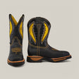 The Rhino Black Composite Toe - E6087 is a pair of black cowboy boots featuring brown soles, intricate yellow stitching, and embroidery on the shafts, reflecting the durability and comfort of Texas Country Work Boots. One boot faces forward while the other shows a side view.