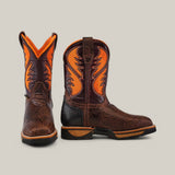 The Jurasic Brown Soft Toe - E6089 cowboy boots, with brown leather and orange flame-like stitching, highlight durability. They have a textured finish, low heels, and pull tabs. Displayed on a plain background, one boot faces forward while the other is turned to the side.