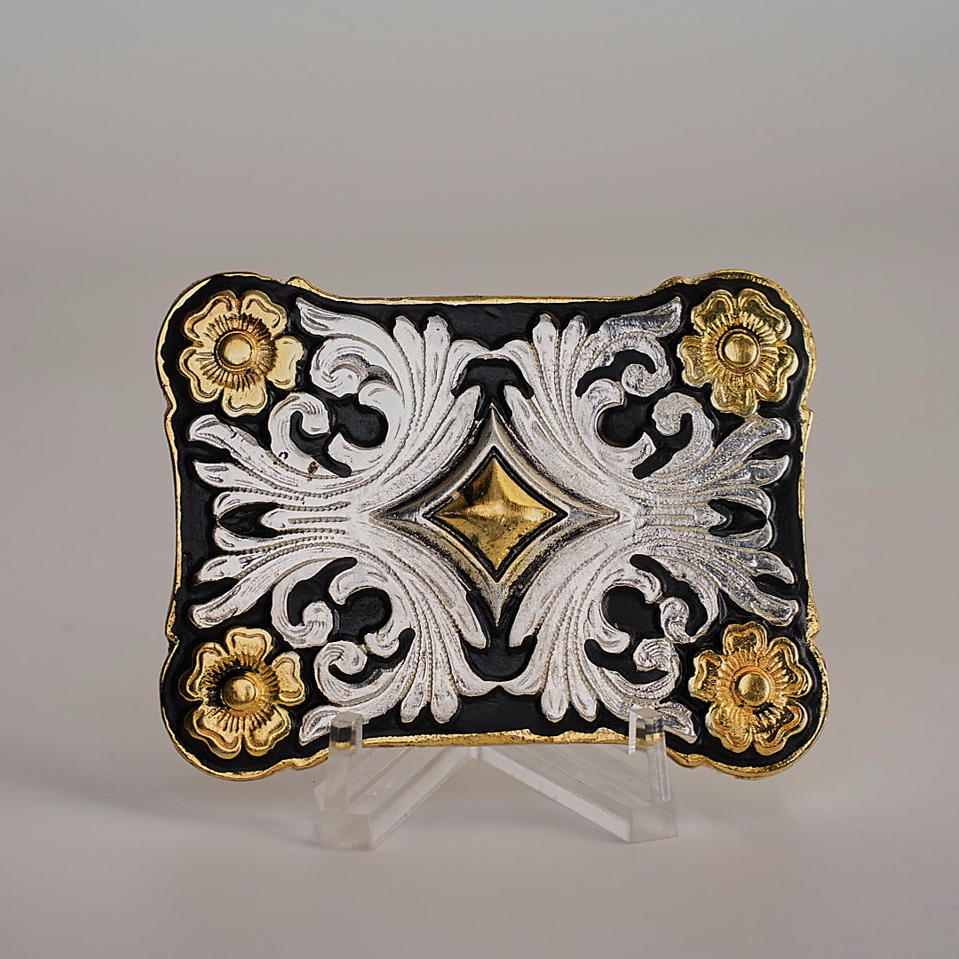 The Floral Diamond Gold Buckle is an ornate plaque with intricate western-style scrollwork, gold floral accents on a black background, and a central raised gold diamond shape, all showcased on a clear stand.