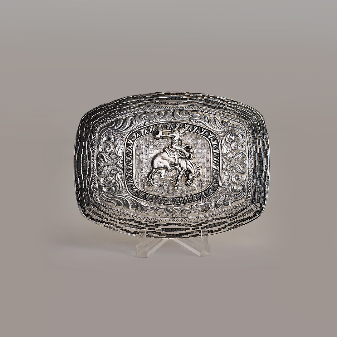 The Charro Floral Bull Rider Buckle, a rectangular Texas-style belt buckle made from alpaca silver, showcases an intricate design with a bull rider at its center, surrounded by ornate floral and scroll patterns. Displayed on a clear stand against a plain background, it exudes Western charm.