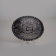 The JGL Buckle, an intricately designed oval piece made from alpaca silver, showcases bold JGL letters and ornate swirling patterns like Texas-style buckles. It is displayed on a clear stand against a plain backdrop.