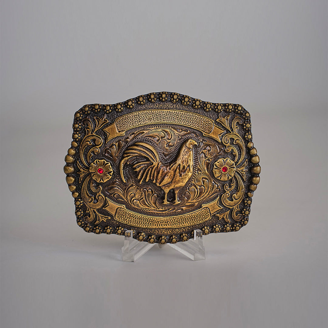 The Ruby Rooster Buckle is an antique brass belt buckle featuring intricate engravings in a classic western design. It showcases a raised rooster centerpiece encircled by floral patterns and embellished with two red gemstone accents, reminiscent of Texas-style buckles. Displayed elegantly on a clear stand.