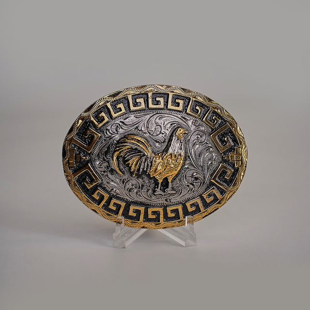 The Charro Rooster Buckle is an oval belt buckle with a western design, featuring a gold rooster on a silver background with swirling patterns. It has a Greek key motif in gold and black on the border. Made from alpaca silver, it is displayed on a clear stand against a gray background.