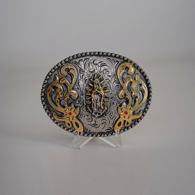 The Virgen Maria Buckle is an ornate oval-shaped belt buckle with intricate western designs, detailed engravings, a central figure, and floral patterns. It features gold accents and red stones, displayed on a clear stand against a neutral background.