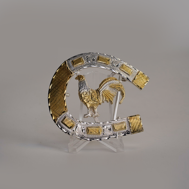 The HorseShoe Rooster is a decorative silver and gold horseshoe with ornate detailing, showcasing a golden rooster centrally. It features intricate patterns and textures, reminiscent of western designs, and is displayed on a transparent stand against a plain background.