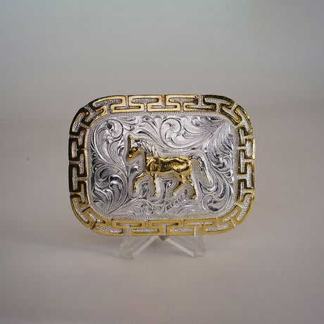 The Charro Full Horse Buckle showcases a rectangular alpaca silver design with intricate western engravings, a central gold horse motif, and golden geometric borders. Elegantly presented on a clear stand, it embodies classic Texas-style craftsmanship.