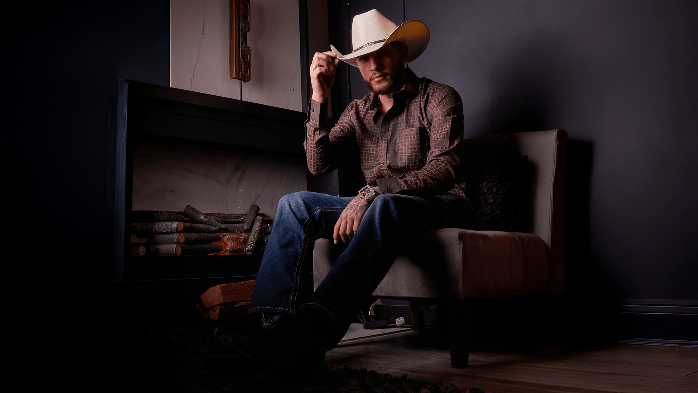 Man dressed in Western attire, wearing a white cowboy hat, brown patterned shirt, blue jeans, and boots, sitting on a beige armchair next to a fireplace in a dimly lit room