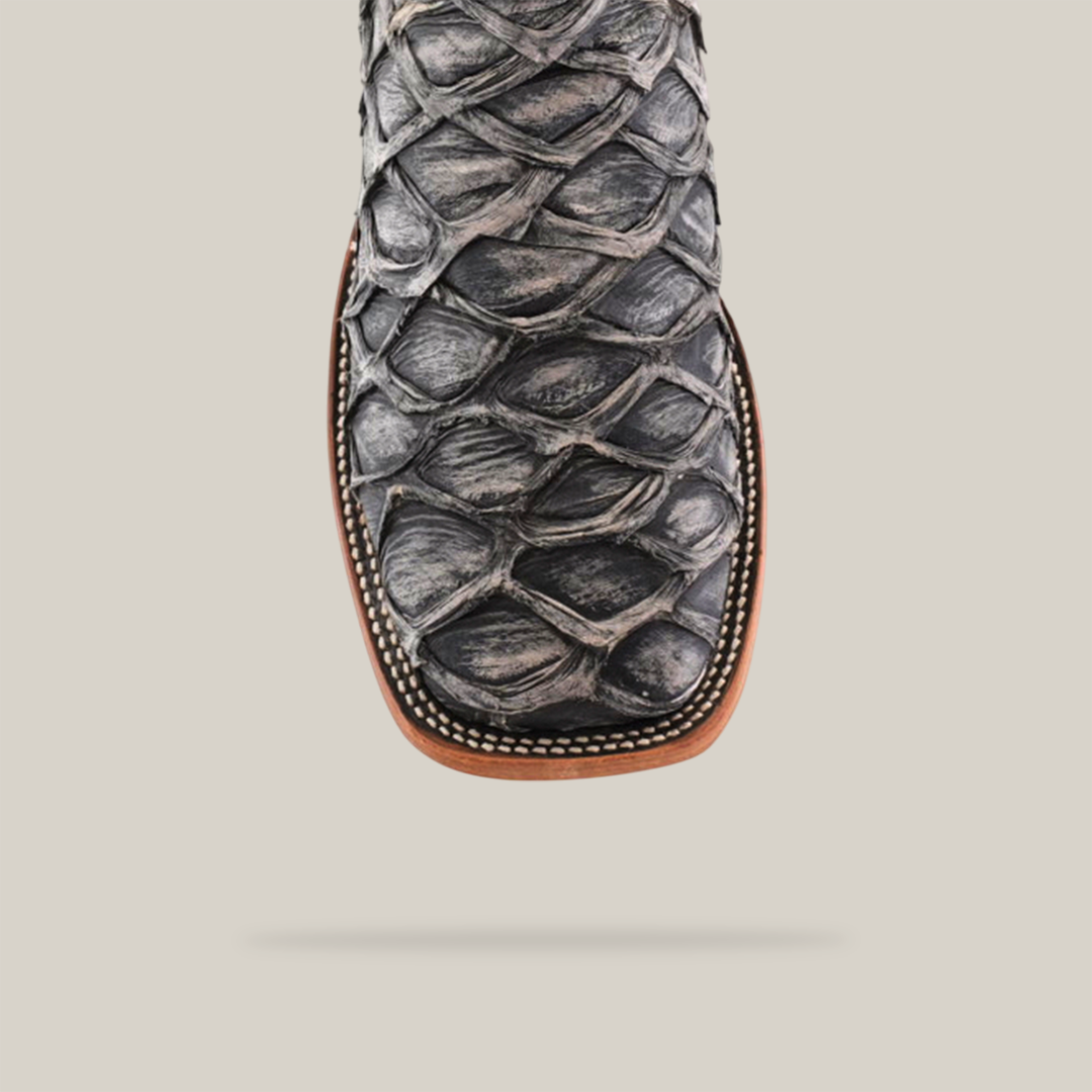 Close-up of the Exotic Piraruco Fish boot, highlighting its textured scaly design and dark leather. The square toe features rustic bone stitching on the edge, set against a light beige background.