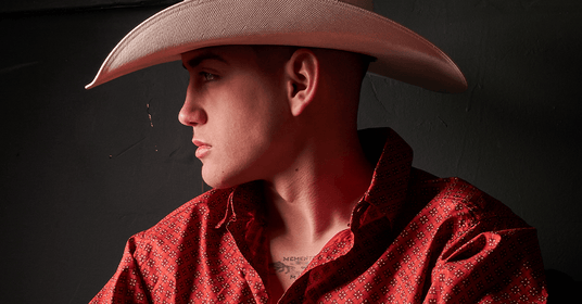 Close-up portrait of a young man in a white cowboy hat and a red patterned Western shirt, gazing to the side against a dark background