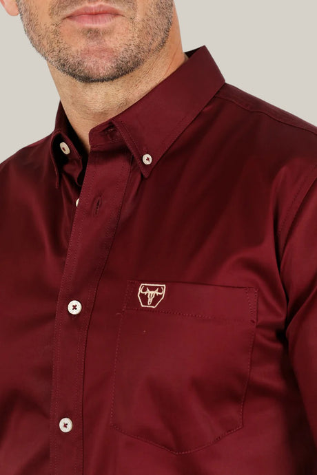 A person is wearing the Men’s Single Pocket Logo Modern Fit Stretch Dress Shirt in Burgundy (CTL9260), featuring a maroon button-up design with a small embroidered bull logo on the breast pocket and a collar, with their face partially visible.