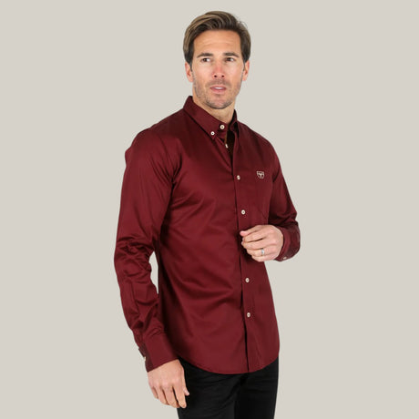 A man in a burgundy Men’s Single Pocket Logo Modern Fit Stretch Dress Shirt (CTL9260) stands against a plain background, looking slightly right while adjusting the buttons.