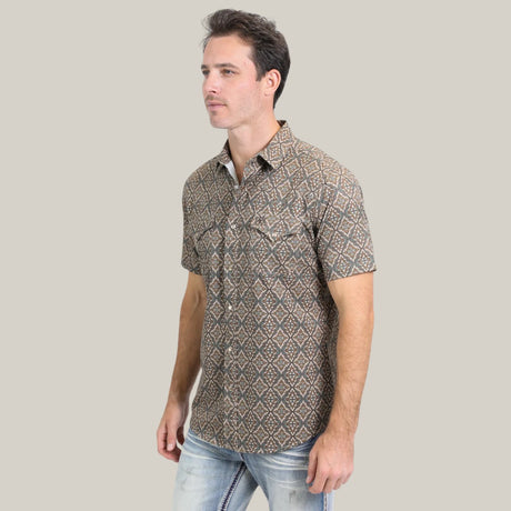 A man in the Mens Performance Western Short Sleeve Aztec Print Brown Shirt - PGW10638 and light blue jeans stands against a plain beige background, slightly facing to the side with short hair and a neutral expression.