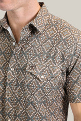 Close-up of someone wearing the Mens Performance Western Short Sleeve Aztec Print Brown Shirt - PGW10638, showcasing brown and teal geometric designs with pearl snap buttons and a chest pocket. The background is neutral, and their face is not visible.