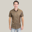 Someone is wearing the Mens Performance Western Short Sleeve Aztec Print Camel Shirt - PGW10613. The brown button-up shirt features a blue geometric Aztec pattern, two chest pockets, and pairs stylishly with light blue jeans against a plain light gray background.