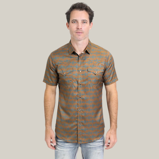 Someone is wearing the Mens Performance Western Short Sleeve Aztec Print Camel Shirt - PGW10613. The brown button-up shirt features a blue geometric Aztec pattern, two chest pockets, and pairs stylishly with light blue jeans against a plain light gray background.