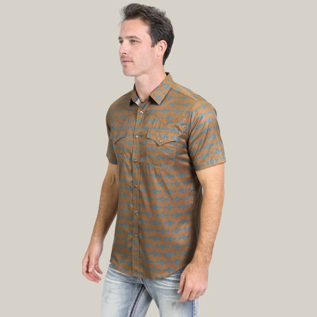 A man wearing the Mens Performance Western Short Sleeve Aztec Print Camel Shirt (PGW10613), featuring a subtle blue geometric Aztec design, paired with light blue jeans, poses against a plain beige background.