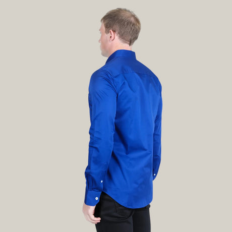 A man wearing the Men’s Single Pocket Logo Modern Fit Stretch Dress Shirt in Royal Blue (CTL9830) pairs it with black pants and stands against a plain, light gray background.