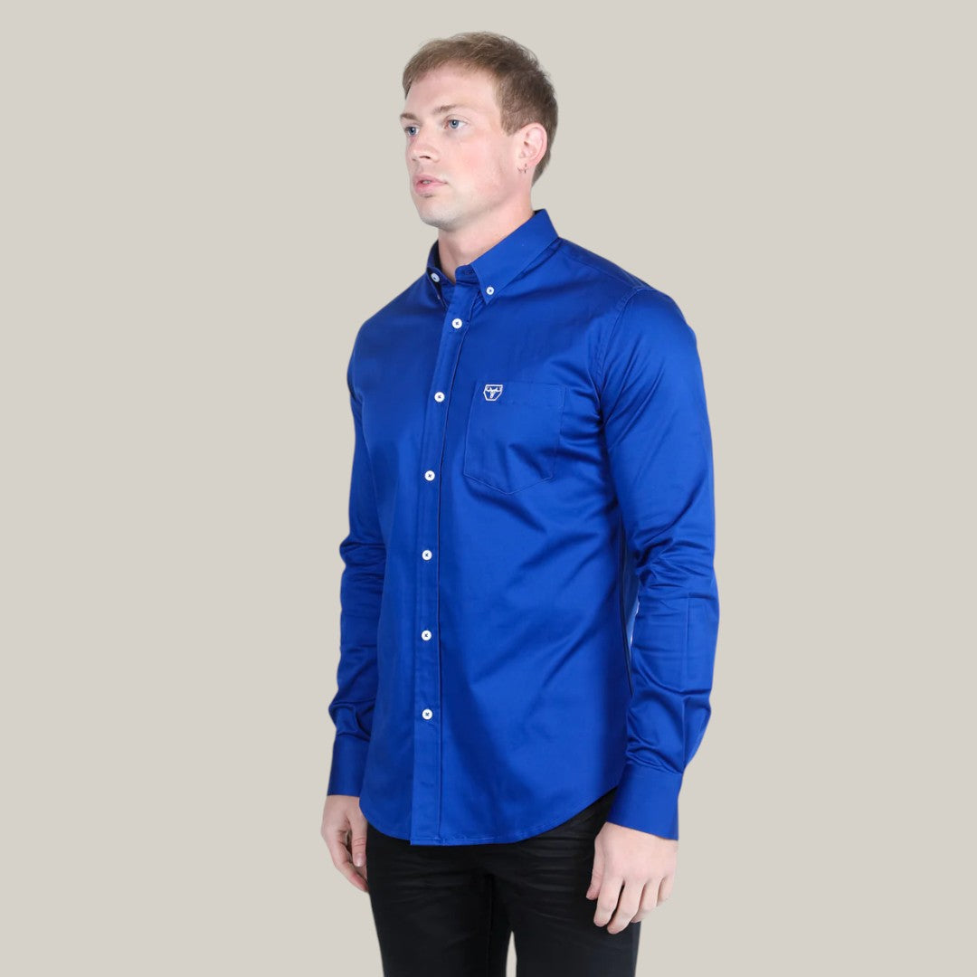 A person wears a Men’s Single Pocket Logo Modern Fit Stretch Dress Shirt in Royal Blue (CTL9830), featuring long sleeves and an embroidered chest logo, against a plain gray background. With a neutral expression, they face slightly left, enjoying comfort from the stretch fabric.