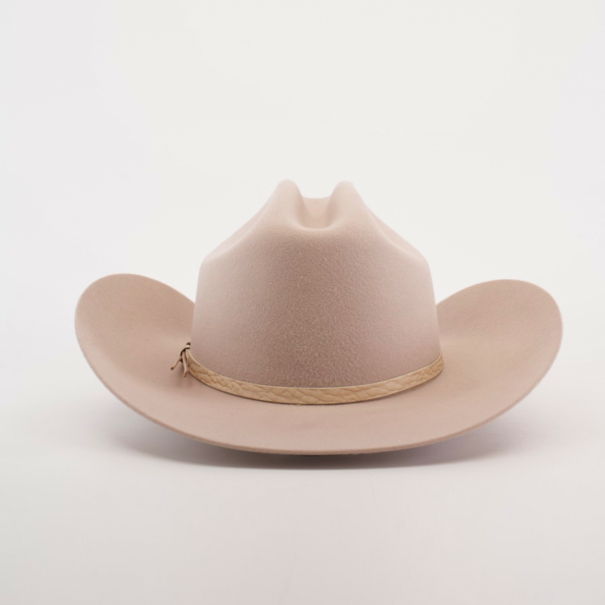 The 6X Country Belly, a beige cowboy hat made from premium sheep wool, features a wide brim and crown crease on a white backdrop. A light brown braided band adds rustic charm to this classic felt hat.