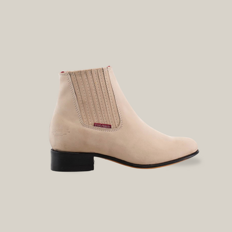 The Prime Suede Pearl Round Toe is a beige suede ankle boot with a low black heel and elastic side panel, featuring textured detail and a red tag. Handcrafted for sleek minimalism on a plain white background, it echoes the timeless style appreciated by the cowboy community.