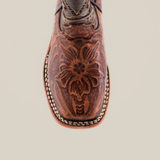 The image showcases a top view of the Handtooled Leave Brown Square Toe boot, crafted from premium cowhide leather. It features an intricate floral carving on the toe and a white and brown stitched edge, enhancing its handmade charm.