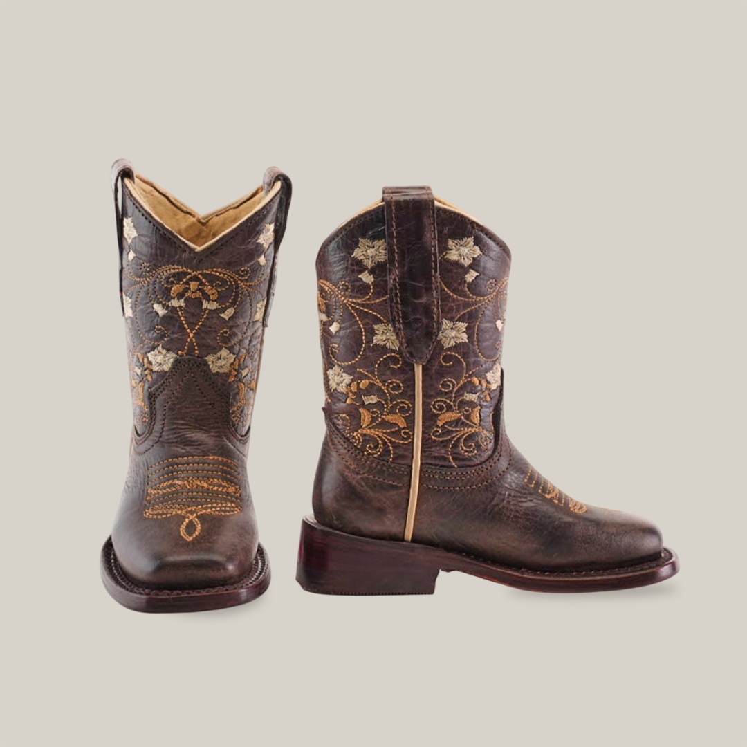 The Dariela Chocolate boots for girls boast premium leather with a square toe and intricate gold floral embroidery. One boot stands upright to showcase the detailed design, while the other is angled slightly on a plain white background.