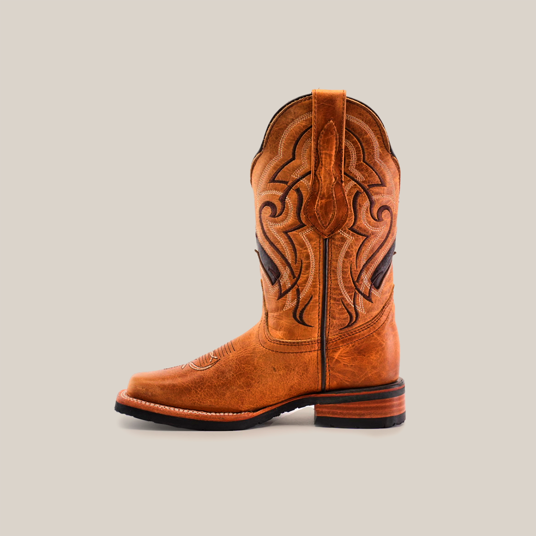 The Wild Mustard Slip Resistant Sole Rodeo Toe boot features a tan cowhide leather design with intricate shaft stitching, a rounded toe, and low stacked heel. It includes pull loops at the top for easy access and offers a slip-resistant sole for stability. Displayed on a white background.