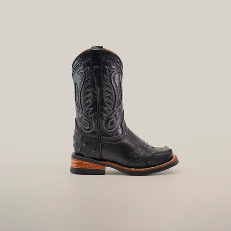 The Karoo Ostrich Half Cowhide Print Black Rodeo Toe boot, featuring intricate stitching on ostrich leather, is set against a white background. It has a wooden heel and textured details on the foot and shaft, offering a classic western style.