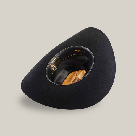 The 6X Real Black hat captures the essence of Western headwear with its asymmetrical shape, rounded crown, and smooth texture. Its interior lining reveals a silky, dark fabric with decorative accents, exuding luxury akin to a Larry Mahan hat.