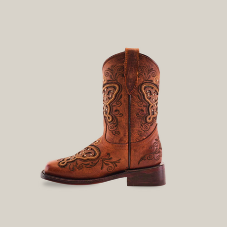 The Butterfly Studs Cogñac boot features a single brown cowhide leather design with intricate embroidery, butterfly studs, swirls, and floral patterns. It has a rounded toe, pull strap, low wooden heel, all displayed against a plain white background.