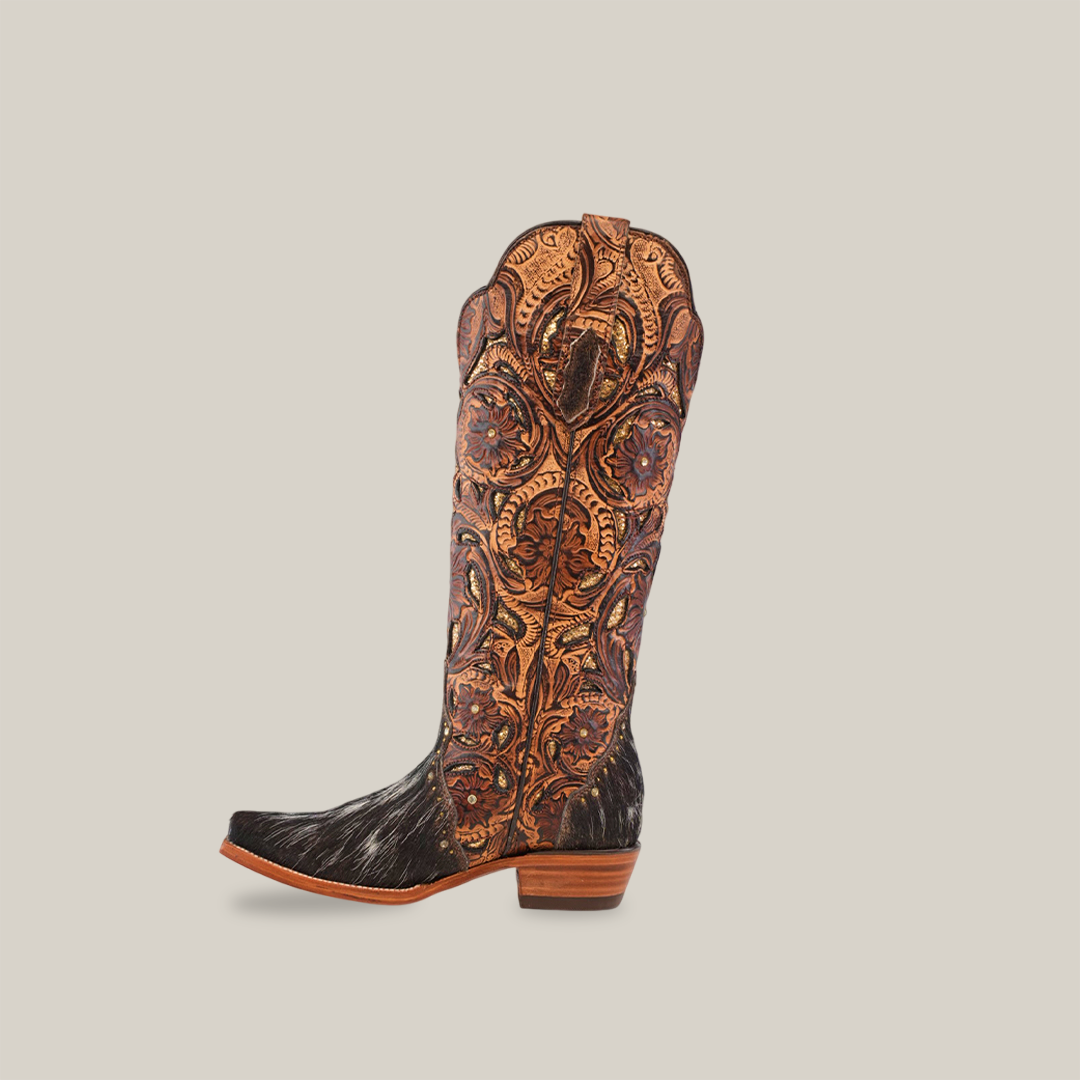 The Handtooled Cowhide Pinto boot is a tall, handcrafted womens boot with intricate brown and orange stitching on the shaft and a black textured cowhide lower, all set against a white background.