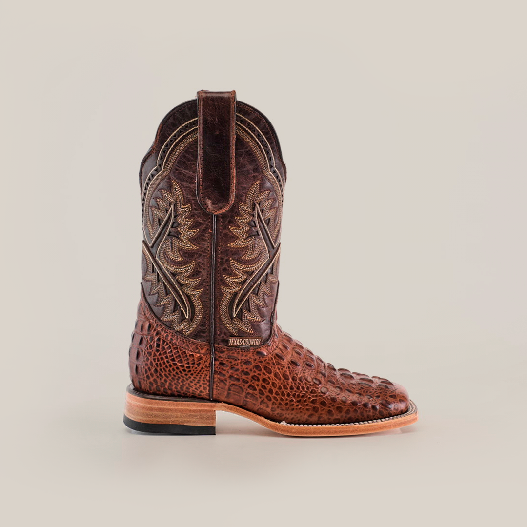 The Caiman Hornback Print Chocolate Square Toe is a single brown cowboy boot crafted from textured leather, showcasing intricate stitching on the upper part. It features a chocolate square toe, sturdy heel, top pull tab, and a contrasting lighter sole that embodies luxury elegance.