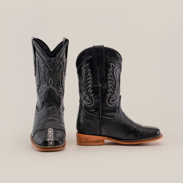 The Stingray Half Cowhide Print Black Rodeo Toe boots exude western charm with intricate stitching and snake-like textured accents. Crafted from cowhide leather, they feature a high shaft and wooden soles, all set against a plain white background.