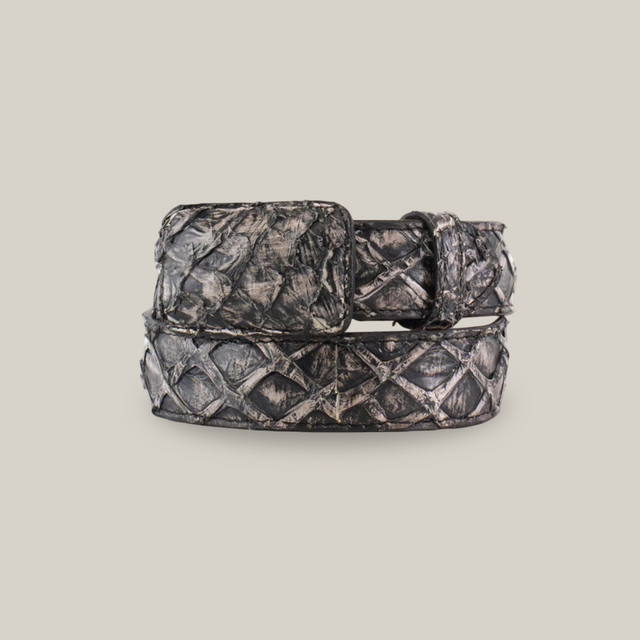 The Exotic Full Quilt Rustic Bone Belt is coiled in a circle, showcasing a diamond pattern with a rectangular buckle. Crafted from Pirarucu fish leather, it has an exotic allure with dark tones and shiny finish resembling metal or leather on a plain white background.