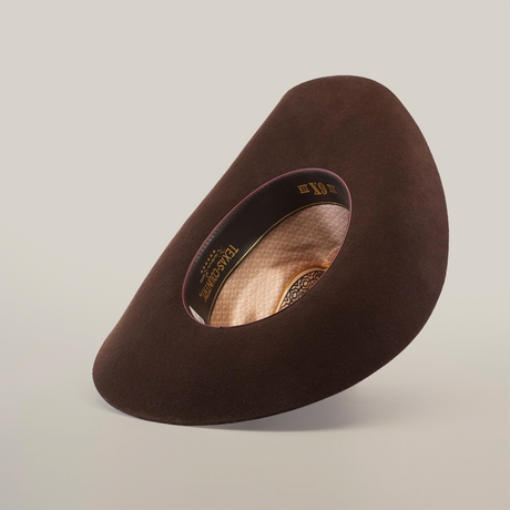 The 6X Country Brown hat, a wide-brimmed felt creation from premium sheep wool, is artfully displayed. Its interior features a patterned lining with TEXANA and XX, embodying Western classic style. The display tilts to reveal both exterior and interior details against a plain background