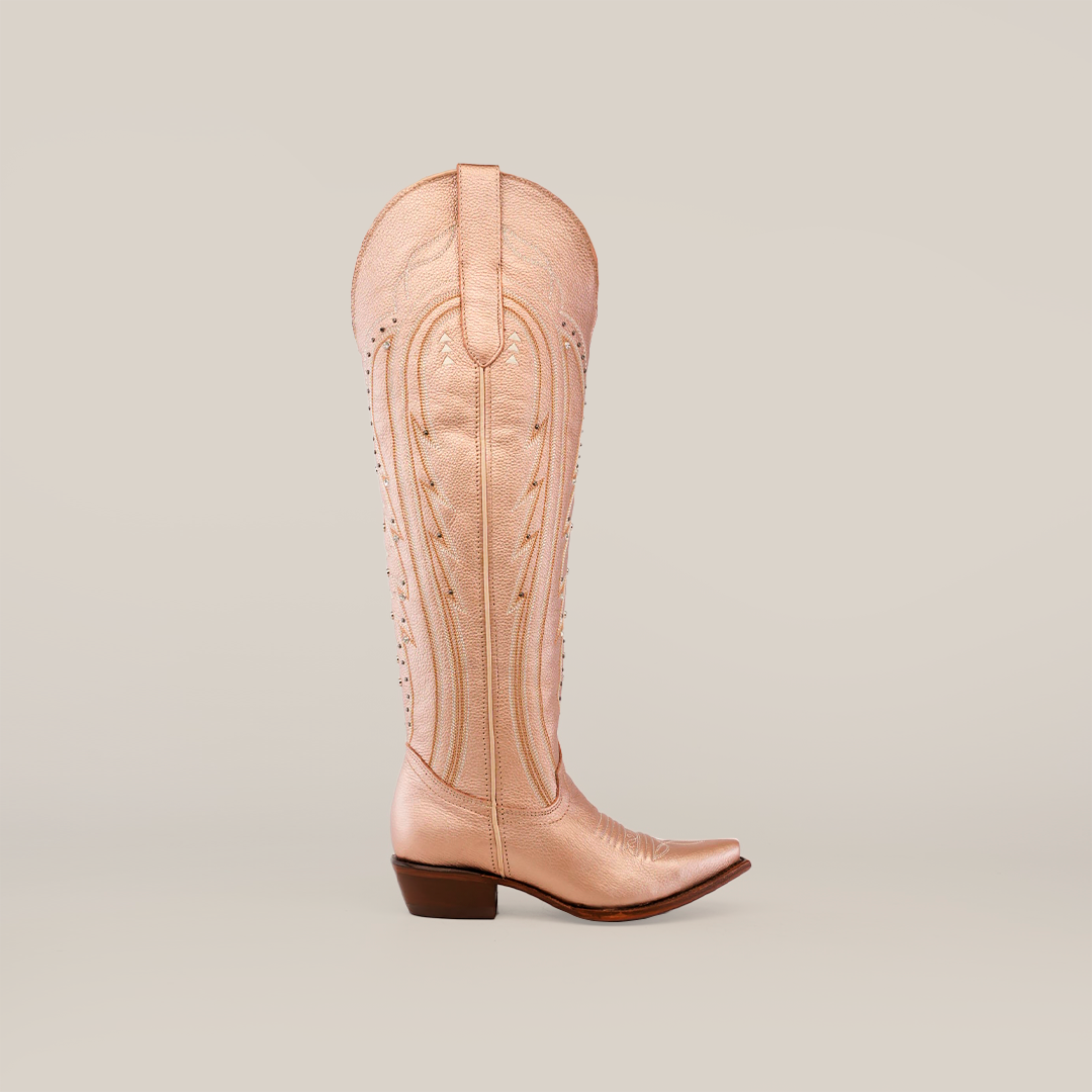 The Sara Metalic Florentine boot, with its tall shaft and snip toe, stands upright against a white background. Its beige color and decorative stitching capture the elegance of classic Western style, featuring a pointed toe and low angled heel.