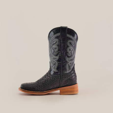 The Python Print Matte Black Square Toe boot is crafted from cowhide leather, featuring a textured surface and intricate white stitching on the upper part, resting on a wooden heel, all set against a plain white background.