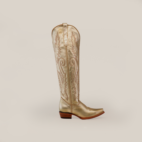 The Brittany Gold - Tall Shaft - Snip Toe womens boot is a metallic gold handcrafted masterpiece featuring intricate stitching and a low wooden heel. Set against a plain gray background, its pointed toe and embroidered shaft pattern make it a standout piece.